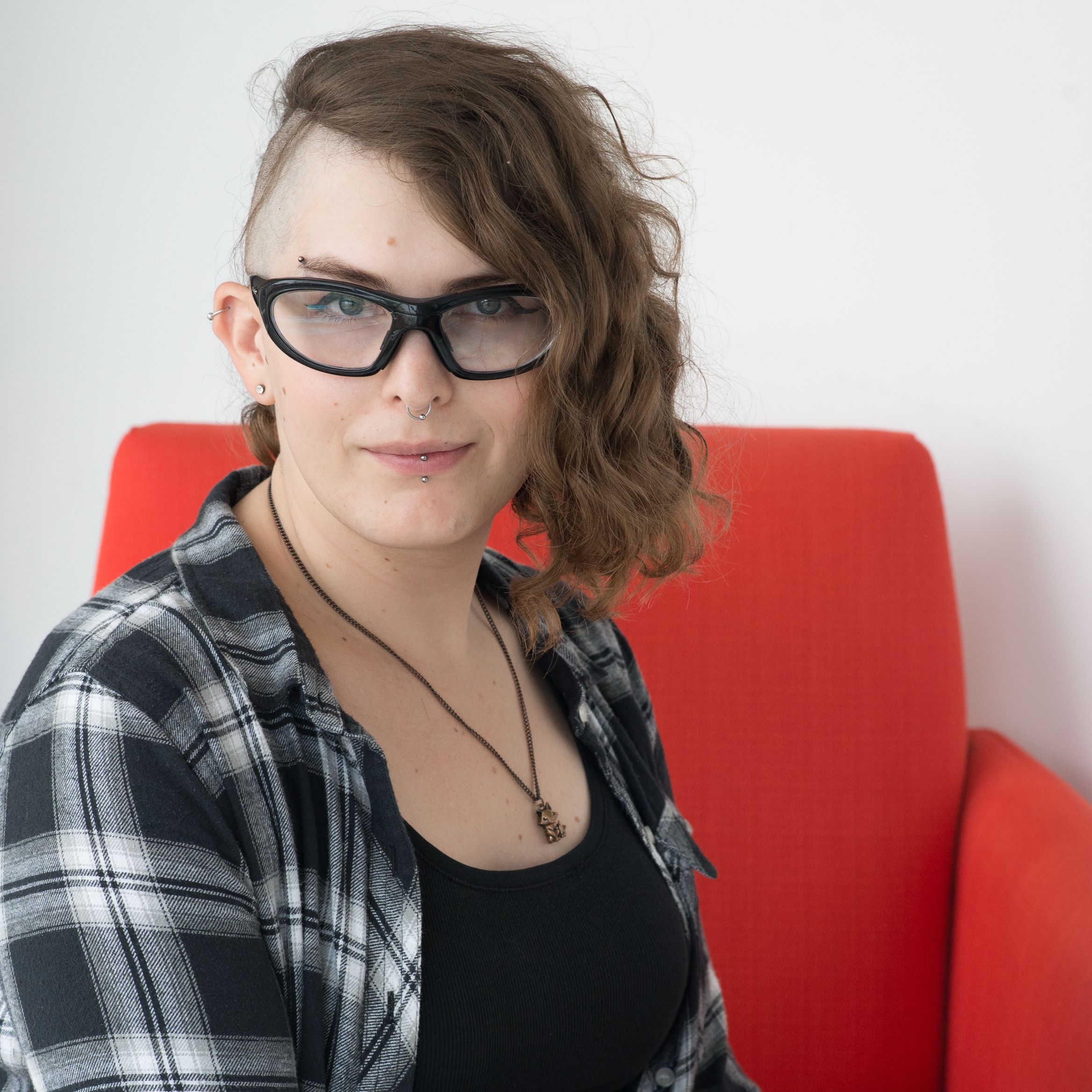 A headshot of June Epstein. She is wearing a black and white plaid shirt and is sitting on a red chair.
